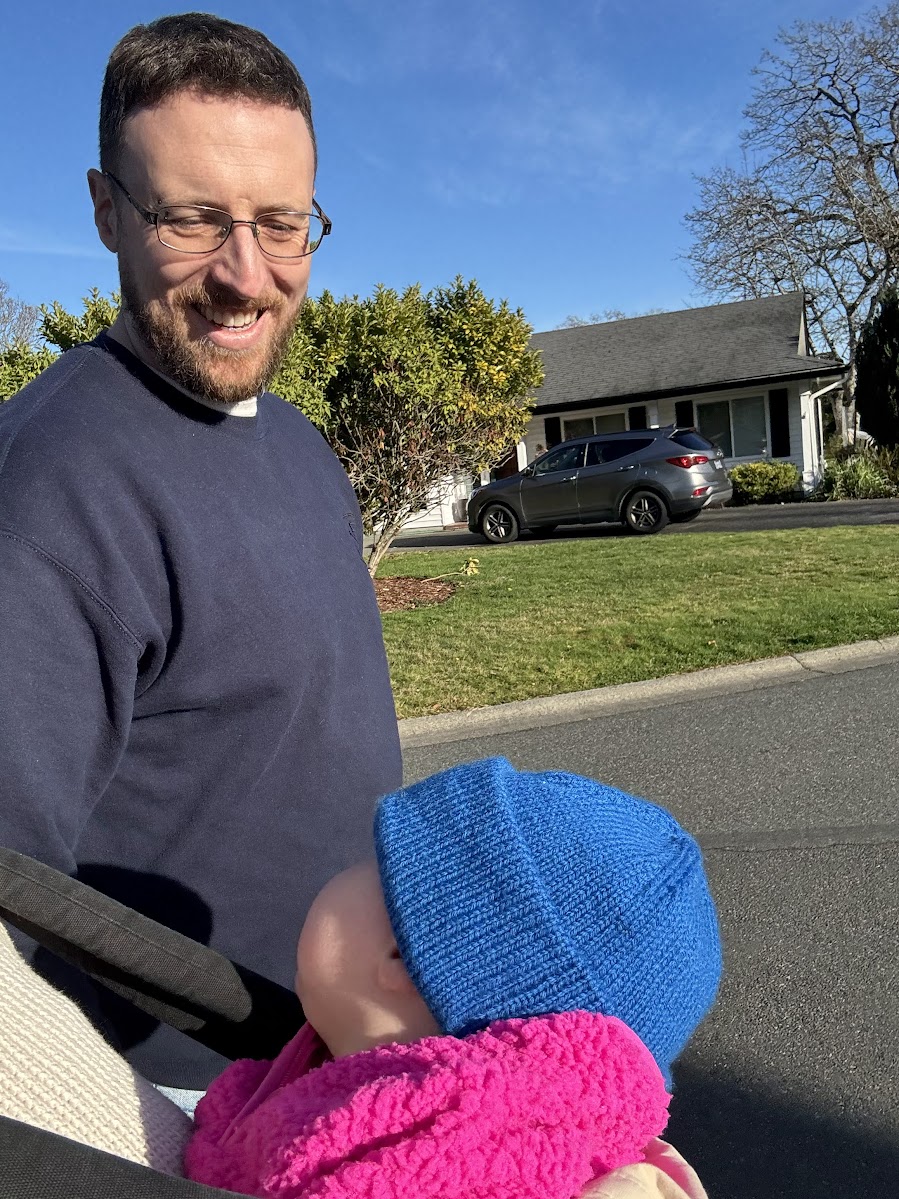 Nick Houghton smiling, holding his baby in a carrier on a sunny street in Victoria, B.C.